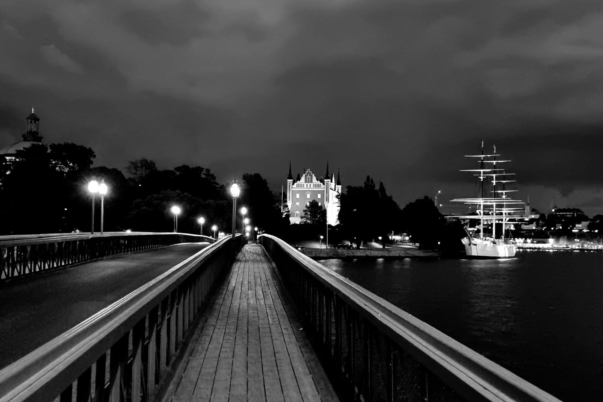Gångväg på en bro med lysande lyktor, omgiven av vatten med ett segelfartyg